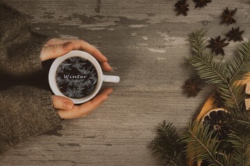 Christmas atmospheric set. Female hands in a cozy green sweaters sleeves with coffee cup and frosty lettering in it. Spruce twigs, anise on a wooden table.