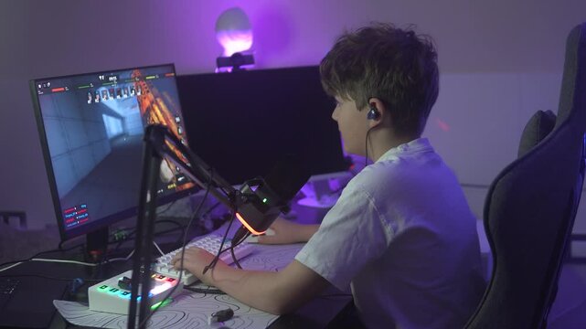 A teenage boy plays a first-person shooter game on a PC while streaming or recording his gameplay. He sits at a gaming setup with a microphone, LED lighting, headphones, and a large monitor. The
