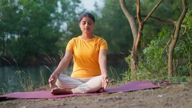 A woman in a yellow shirt meditates on a yoga mat by a calm lake, surrounded by trees and nature.
