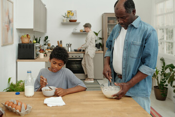 African American father preparing meal in kitchen while son eating cereal, mature woman cooking in...