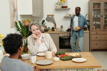 Caucasian woman sitting at dining table sharing breakfast with multiethnic family in cozy kitchen setting. Man in background bringing dishes, creating relaxed atmosphere