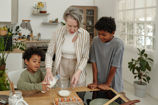 Woman guiding two children in baking activity inside sleek kitchen with wooden and metallic elements and bright natural light. Kitchen filled with various cooking ingredients around