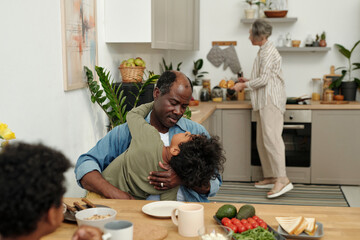 African American father holding child in bright kitchen while older woman prepares food in...