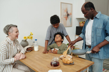 Family gathering and engaging in breakfast preparations around wooden table. Caucasian woman, two children, and senior African American man interacting