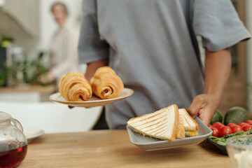 Person holding plates with croissants and sandwiches in cozy kitchen setting. Fresh fruits and...