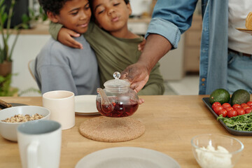 Siblings embracing while adult hand pours tea in cozy kitchen setting with mugs, cereal bowls, and fresh vegetables around. Warm and homely atmosphere