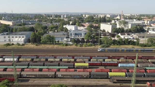 An aerial view of a busy railway station with multiple freight and passenger trains on parallel tracks. The background features an urban landscape with residential and industrial buildings.