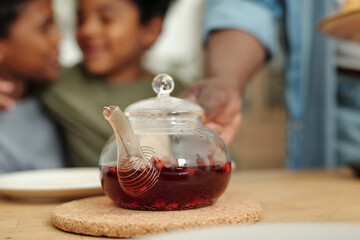 Unknown man putting glass teapot with loose tea leaves on table. Blurred background emphasizes focus on tea preparation process and interaction of two children