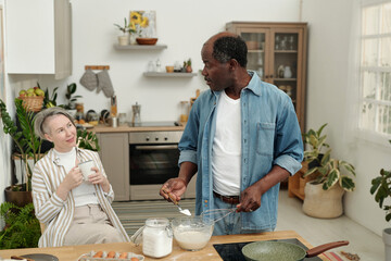 Multiethnic couple preparing breakfast in modern kitchen, surrounded by plants and cooking utensils, sharing moment of conversation and laughter