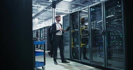 Male Data Center IT Engineer Standing in a Room with Server Racks. Cloud Computing Architect Using Laptop Computer for Servicing the System in Cyber Security and Data Protection Facility - Powered by Adobe