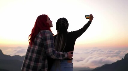 Couple embrace taking selfie atop mountain range overlooking cloudscape during sunset