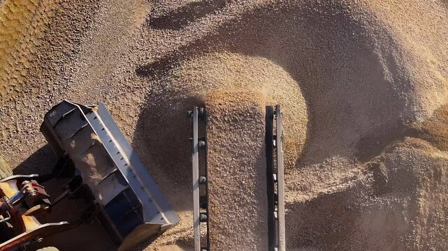 Top-down aerial of a conveyor belt dropping crushed stone onto a pile. A wheel loader works at the quarry, producing construction materials like sand and gravel.
