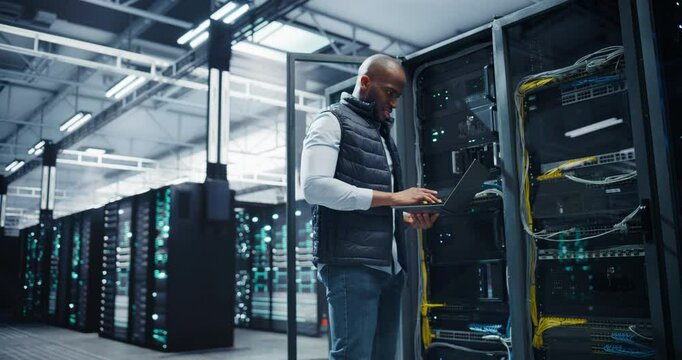 African Server Technician Installing Additional Hardware into Server Cabinet in a Data Center. Specialist Running Diagnostics on a Laptop Computer, Optimizing Storage Hard Drives in a Modern Facility