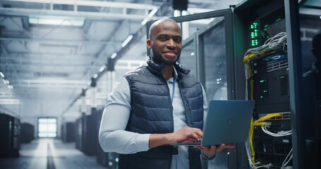 Portrait of a Data Center Engineer Using a Laptop Computer. Young Black Man Looking at Camera and Smiling. Information Technology Specialist Standing in a Server Room Facility at a Research Center - Powered by Adobe