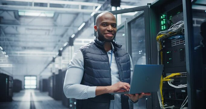 Portrait of a Data Center Engineer Using a Laptop Computer. Young Black Man Looking at Camera and Smiling. Information Technology Specialist Standing in a Server Room Facility at a Research Center - Powered by Adobe
