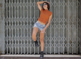 Pretty, chic Asian woman poses stylishly against a metal gate, in a rust crop top, distressed denim shorts, and black combat boots, looking cute with an urban edge