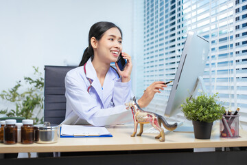 A confident Asian female veterinarian sits at her desk, working on her mobile phone. She manages...