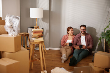 A couple sits on the floor of their new home, enjoying coffee while surrounded by unpacked boxes...