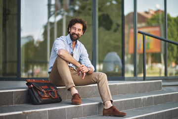 A smiling man dressed in smart casual clothing sits comfortably on outdoor stairs, taking a break with a messenger bag beside him in an urban setting.