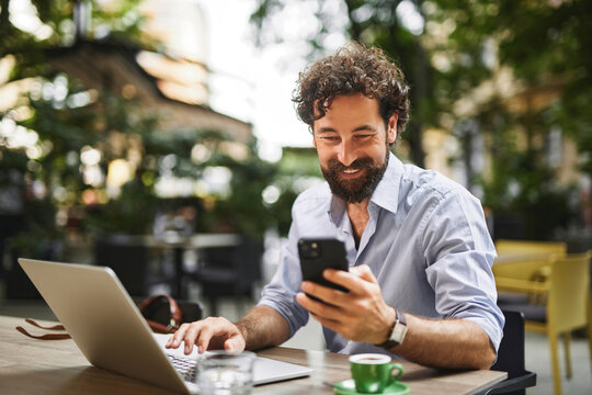 Man sits at a table outside a cafe, using a laptop while looking at his smartphone. He smiles, sipping coffee, surrounded by greenery during a sunny day.