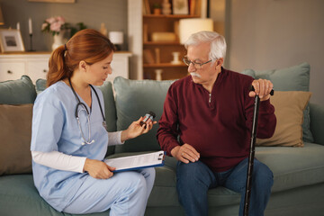 Obraz premium A female caregiver in scrubs reviews medication details with a senior man holding a cane. They are sitting on a couch in a living room during daytime. The nurse holds a clipboard.