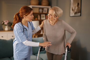 A caring home nurse in scrubs assists an elderly woman using a walker in her living room. The nurse is providing support and encouragement during the daytime in a comfortable home setting.