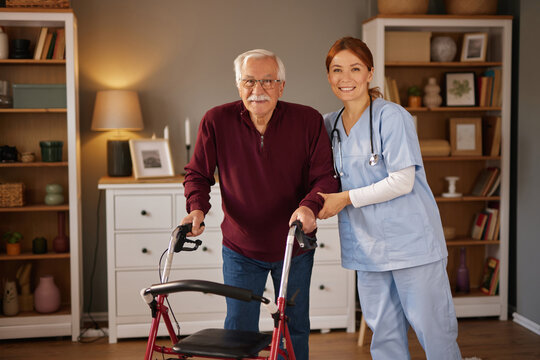 An elderly man uses a walker while a female home healthcare nurse assists him. The scene takes place in a well lit living room, during a daytime appointment. Both subjects are smiling.