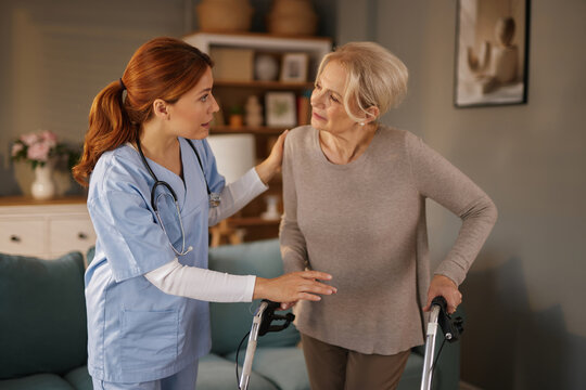 A caring home nurse in scrubs assists an elderly woman using a walker in her living room. The nurse is providing support and encouragement during the daytime in a comfortable home setting.