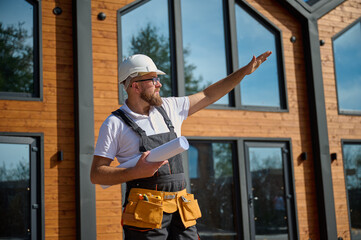 Architect hold plan and pointing on house. Bearded architect wearing hardhat holding project...