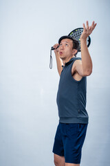 Asian man in sleeveless hoodie and shorts lifts padel racket behind shoulder with one hand raised,...