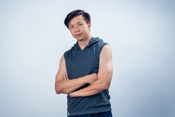 Asian man standing with arms crossed, wearing a dark sleeveless hoodie and navy pants, posing...