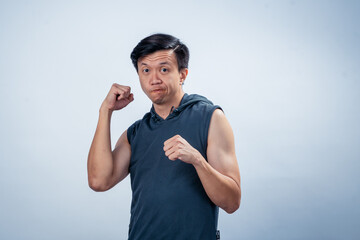 Asian man in sleeveless hoodie showing excited expression while flexing arms and clenching fists,...