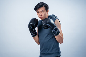 Asian man in sleeveless hoodie wearing boxing gloves strikes forward fighting pose with intense...