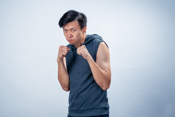 Asian man wearing sleeveless hoodie strikes boxing stance with clenched fists and focused face,...