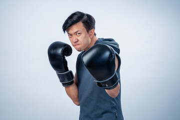 Asian man in sleeveless hoodie wearing boxing gloves strikes forward fighting pose with intense...