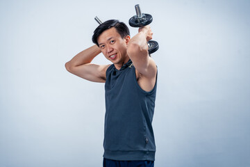 Asian man in sleeveless hoodie lifting dumbbell behind head with both arms, smiling confidently...