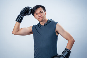 Asian man in sleeveless hoodie wearing boxing gloves presses glove against head with silly facial...