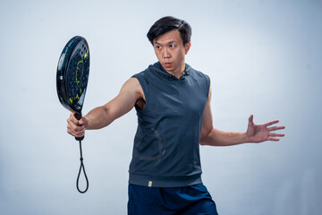 Asian man in sleeveless hoodie holding black padel racket with both hands in mid-swing position,...
