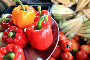 Colorful Bell Peppers and Vegetables at Local Market. A vibrant collection of red and yellow bell peppers on a metal tray surrounded by fresh tomatoes and bundled vegetables.