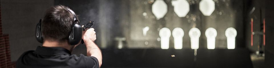 Header of Caucasian middle aged man wearing protective earmuffs practicing shooting with handgun at indoor gun range, aiming at multiple illuminated targets in distance © DragonImages