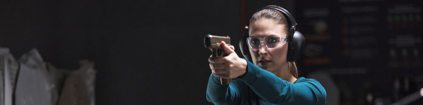 Header of young Caucasian woman wearing protective glasses and earmuffs aiming handgun at shooting range, focusing on target during club training session