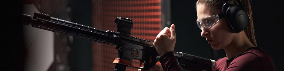 Caucasian young woman wearing protective glasses and earmuffs reloading rifle at indoor shooting club range, focusing on firearm safety and preparation before target practice © DragonImages