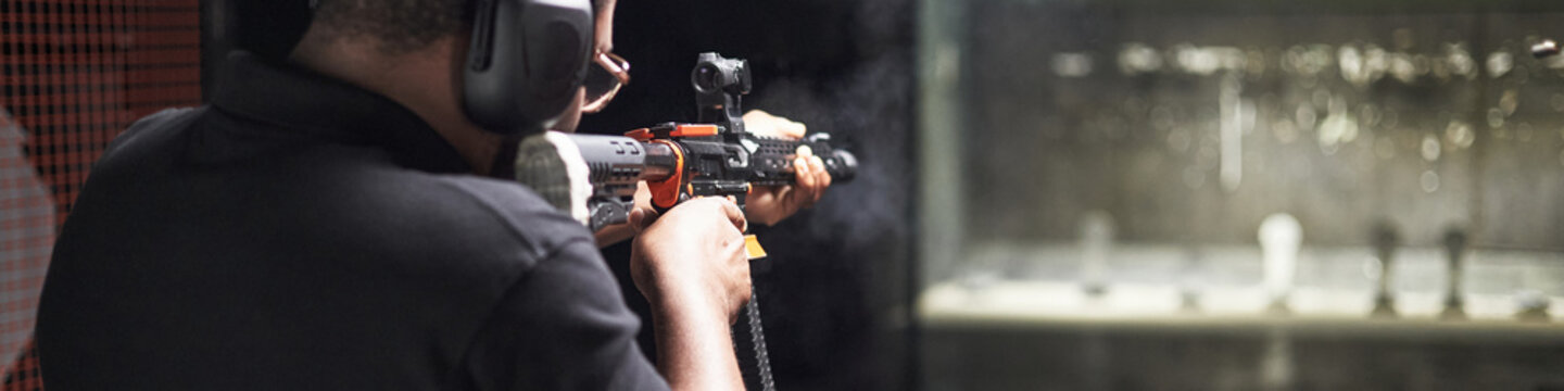 Black man aiming rifle at indoor shooting range, wearing protective earmuffs, focusing on target practice in controlled environment, participating in practice activity