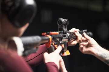 Anonymous female customer wearing protective earmuffs aiming rifle at shooting club while male instructor assisting by adjusting firearm position during training session