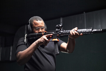 Mature Black male customer aiming rifle at indoor range, wearing protective headphones and safety...