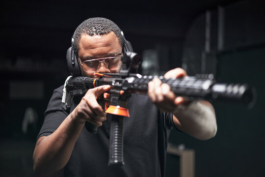 Focused Black man aiming rifle at shooting range, wearing protective glasses and earmuffs, focusing on target during training session, demonstrating firearm handling skills