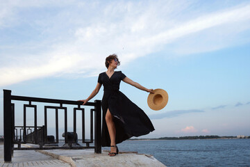 Woman holds straw hat while posing against coastal breeze.