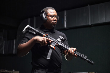 Black middle aged man wearing protective earmuffs holding semi automatic rifle at shooting range,...