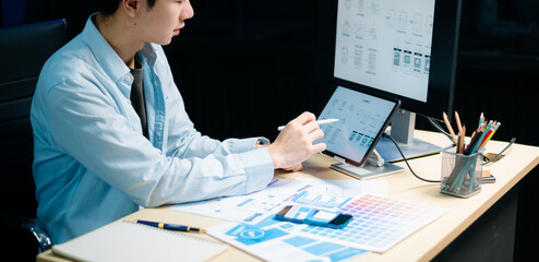 Asian designer’s hands reviewing UX/UI mockups and color palettes at modern desk. App design,...