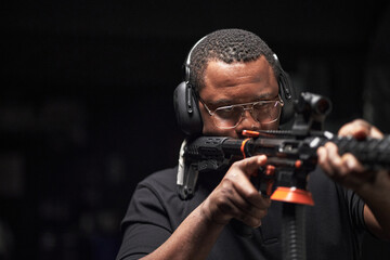 Middle aged Black man wearing protective headphones aiming rifle at shooting range, focusing...